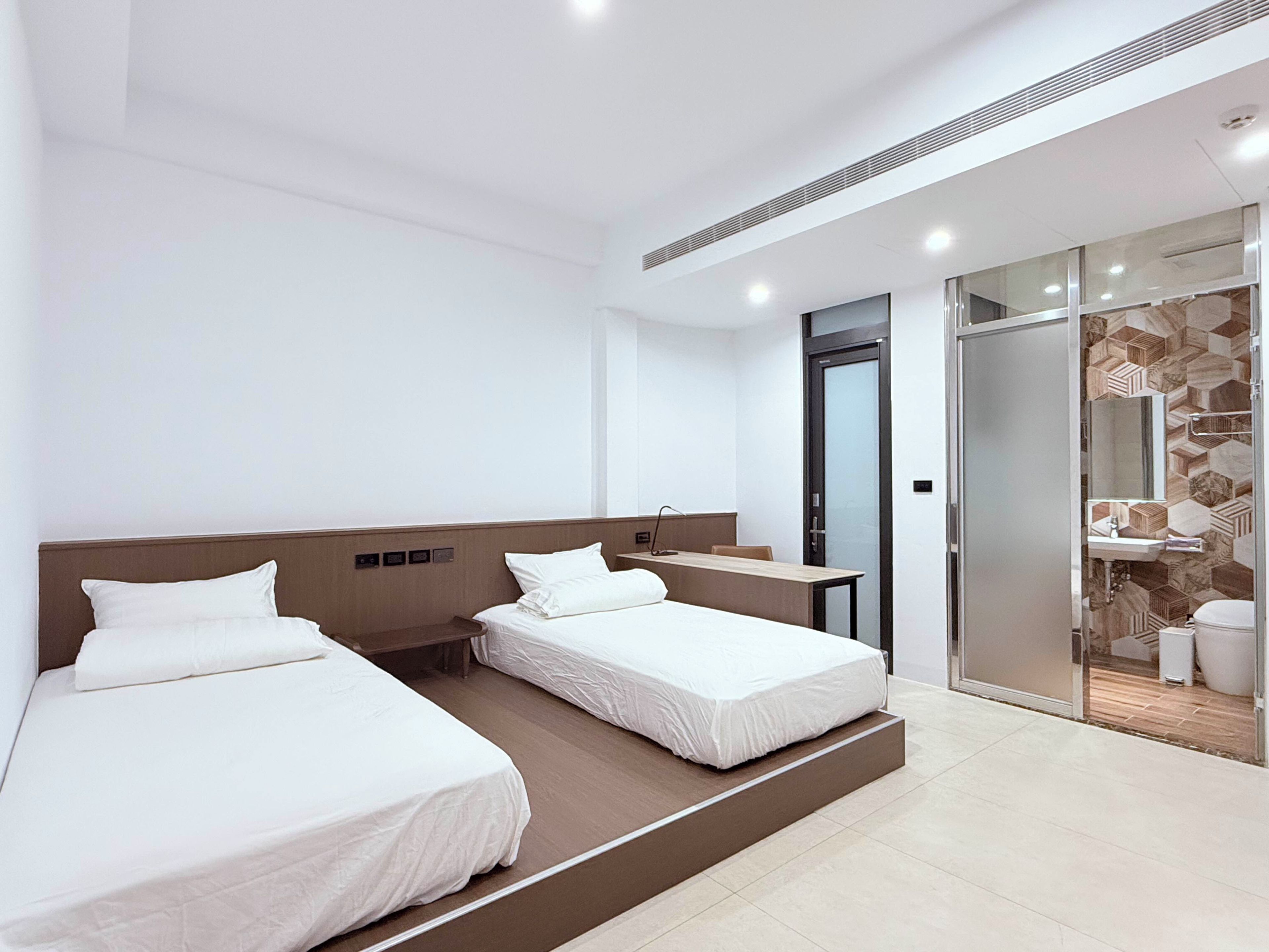A modern hotel room with two beds, a desk, and a bathroom visible through a glass door.