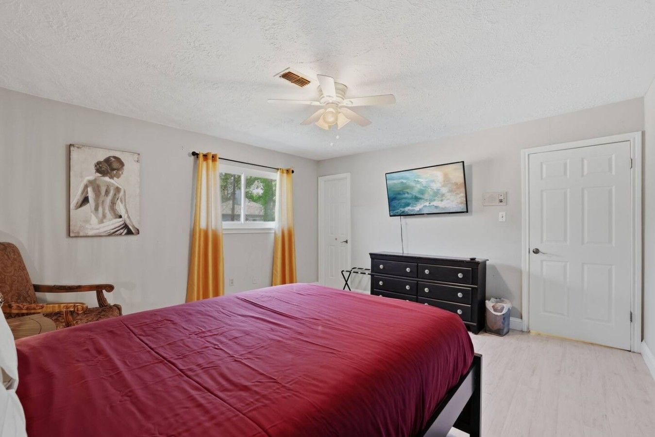 A bedroom with a bed, a chair, a dresser, a ceiling fan, and a painting on the wall.