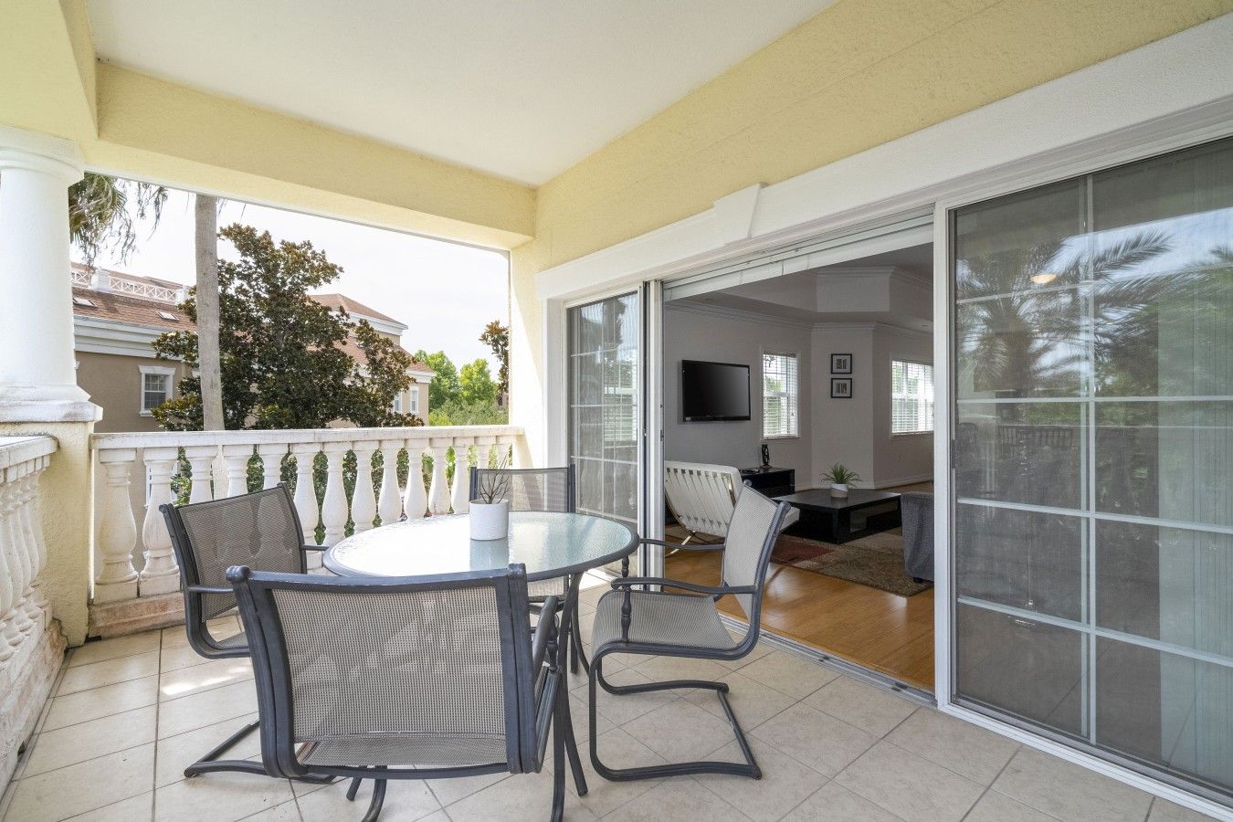 Un patio techado con una mesa redonda y sillas, una puerta corrediza de vidrio que da a un espacio interior con una televisión y una planta.