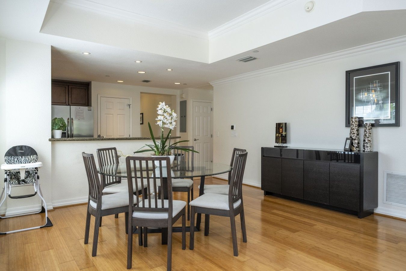 Comedor con mesa de vidrio rodeada de sillas, cocina al fondo y una consola negra con objetos decorativos.