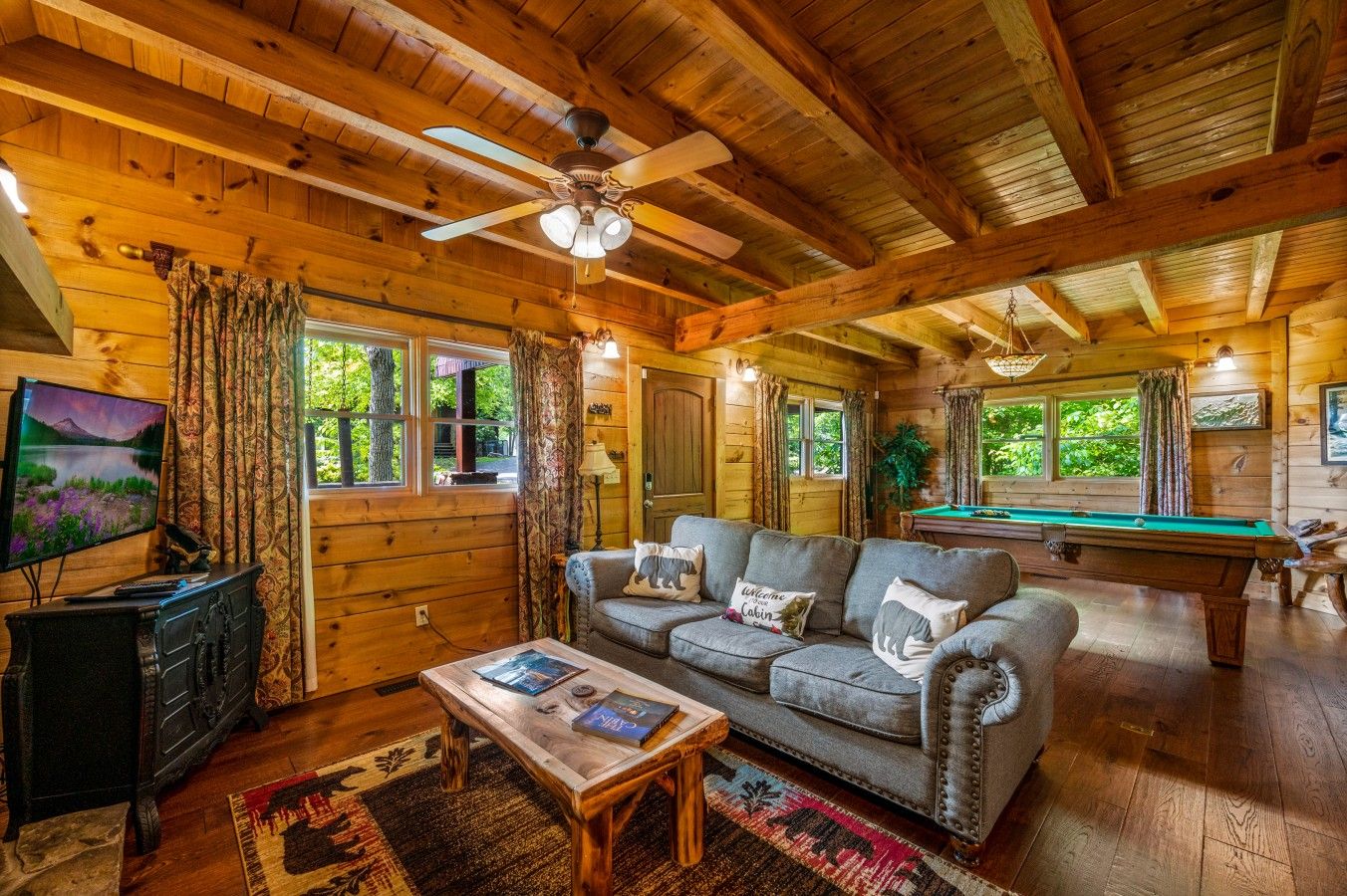 A cozy cabin interior with a wooden ceiling, a sofa, a pool table, and a television.