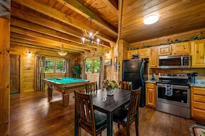 Ein Interieur einer Holzhütte mit Billardtisch, Esstisch, Stühlen, einer Küche mit modernen Geräten sowie einer Decke mit Holzbalken und einem Kronleuchter.