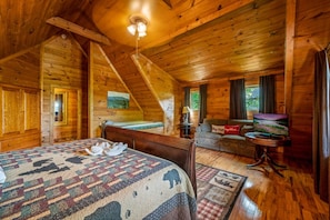 Ein gemütliches Schlafzimmer in einer Blockhütte mit einem großen Bett, einer Couch, einem Fernseher und Blick ins Freie.