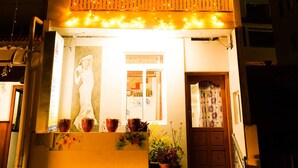 A multi-story building with balconies adorned with string lights, a visible entrance with a closed door, and potted plants at the entrance.