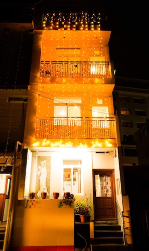 A multi-story building with balconies adorned with string lights, a visible entrance with a closed door, and potted plants at the entrance. - MSP Amma Cottage (Munnar)