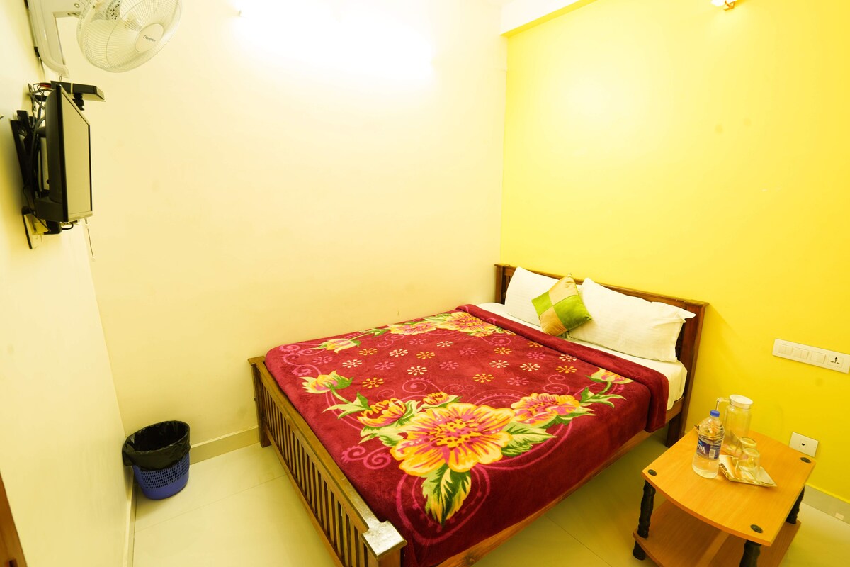 A small, single-bed room with a wooden headboard, a red floral bedspread, a bedside table with a water bottle, and a trash bin.