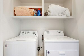 Un lavadero con dos lavadoras Whirlpool blancas y un estante superior con ropa doblada y productos de limpieza.