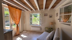 A room with a bed, a window with a view of greenery, a small table with a lamp, and a wall-mounted air conditioning unit.