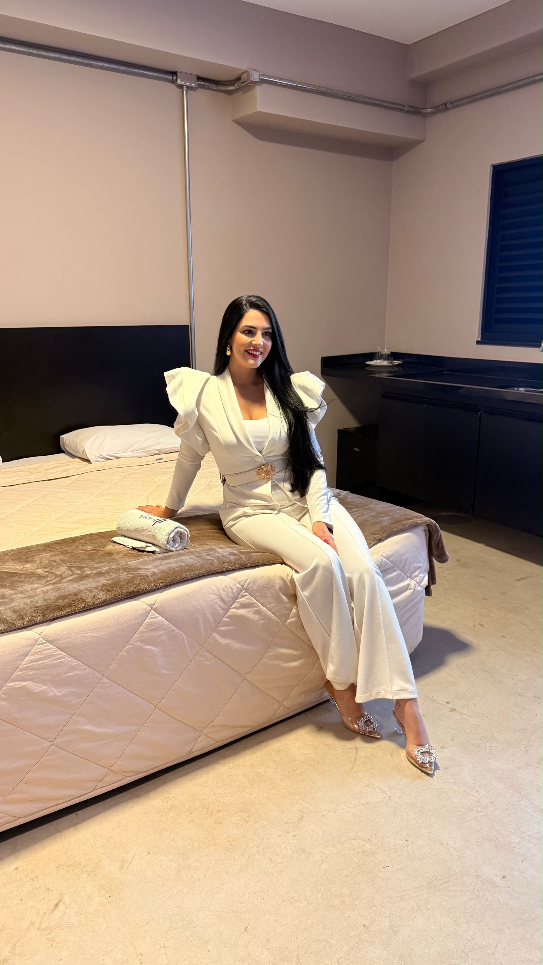 Una persona sentada en una cama en una habitación con cabecera, mesita de noche y cómoda.