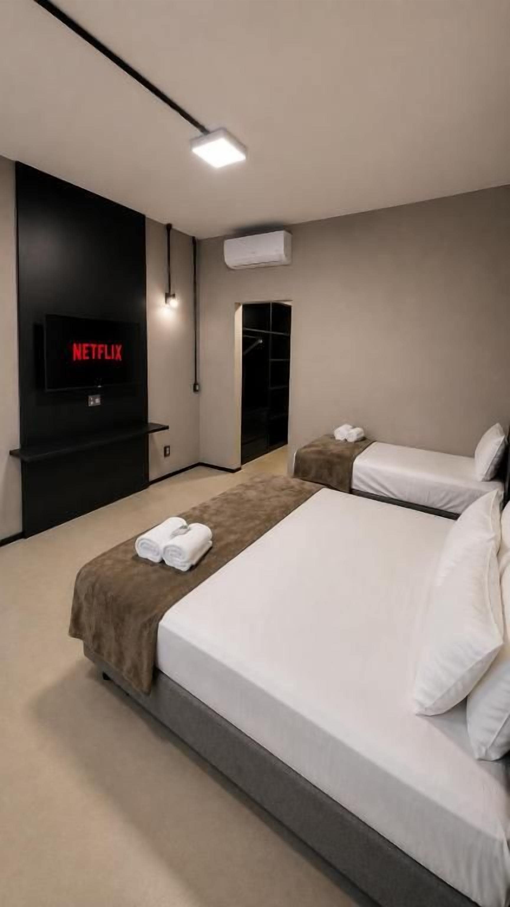 A modern hotel room with a large bed, a TV displaying 'NETFLIX,' and a wall-mounted air conditioning unit.