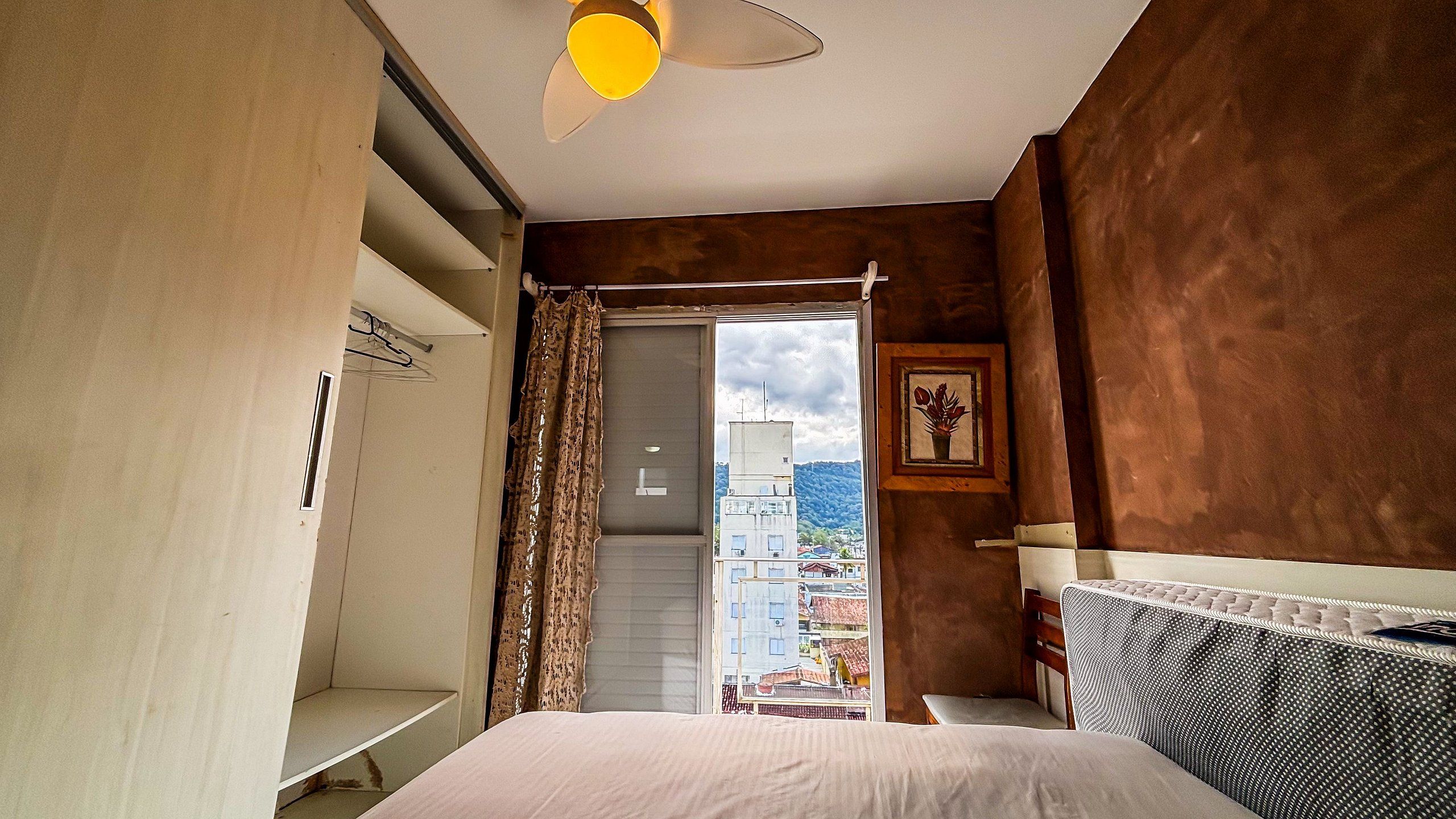Un dormitorio con cama, mesita de noche, ventana con cortinas, lámpara de pared y vistas a edificios.