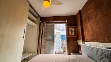 Un dormitorio con cama, mesita de noche, ventana con cortinas, lámpara de pared y vistas a edificios.