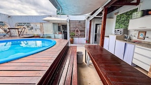 A pool area with a wooden deck, a small trash bin, and a kitchen area with cabinets and a countertop. - SHB Cobertura e Piscina Privativa  (GUARUJÁ)