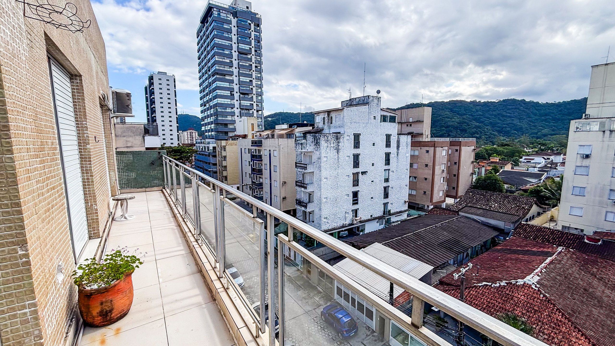 Un balcón con una maceta, un coche estacionado abajo y vistas de edificios y colinas circundantes.