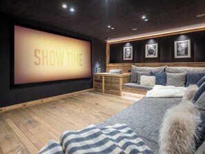 Ein gemütliches Zimmer mit einem großen Bildschirm, der 'SHOW TIME' anzeigt, einer hölzernen Bank, einem Sofa mit weicher Decke und gerahmten Bildern an der Wand.