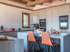 A modern kitchen with a central island, bar stools, a built-in oven, and a window with a view. - Chalet Marianna — Luxury 500m², Private Spa, 180° Mont Blanc Views, Near Megève (Combloux)