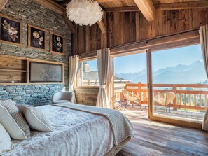 Ein Schlafzimmer mit einem großen Bett, einer Steinwand, Holzbalken an der Decke und einem Balkon mit Bergblick.