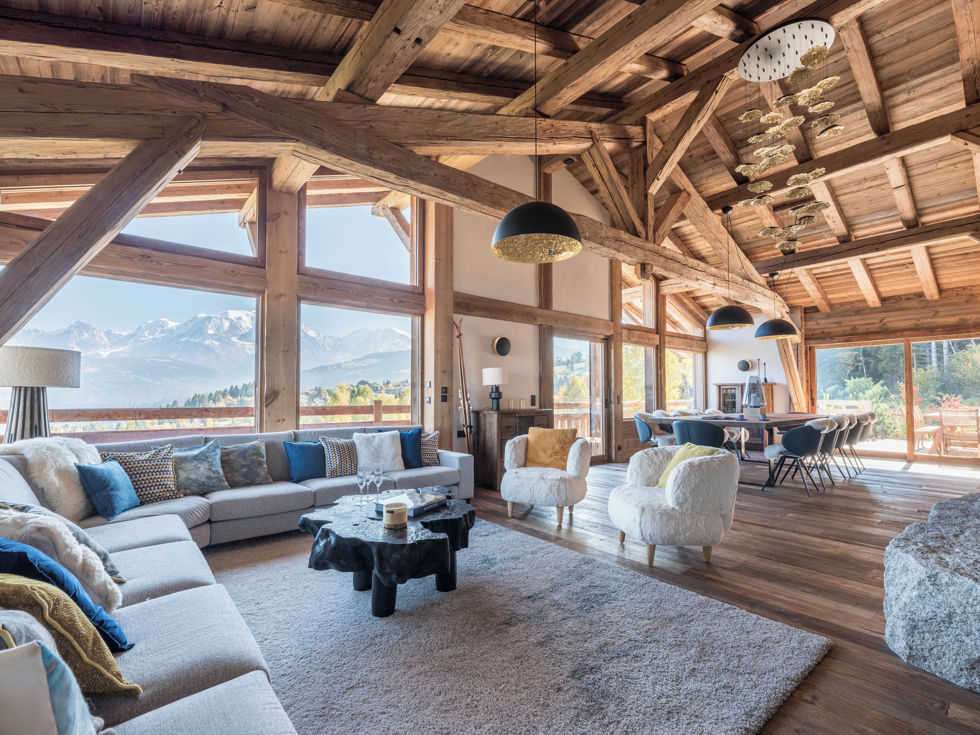 A spacious living area with a large sofa, a coffee table, and a dining table set for four. The room features high ceilings with exposed wooden beams and large windows offering a mountain view.