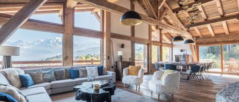 A spacious living area with a large sofa, a coffee table, and a dining table set for four. The room features high ceilings with exposed wooden beams and large windows offering a mountain view.