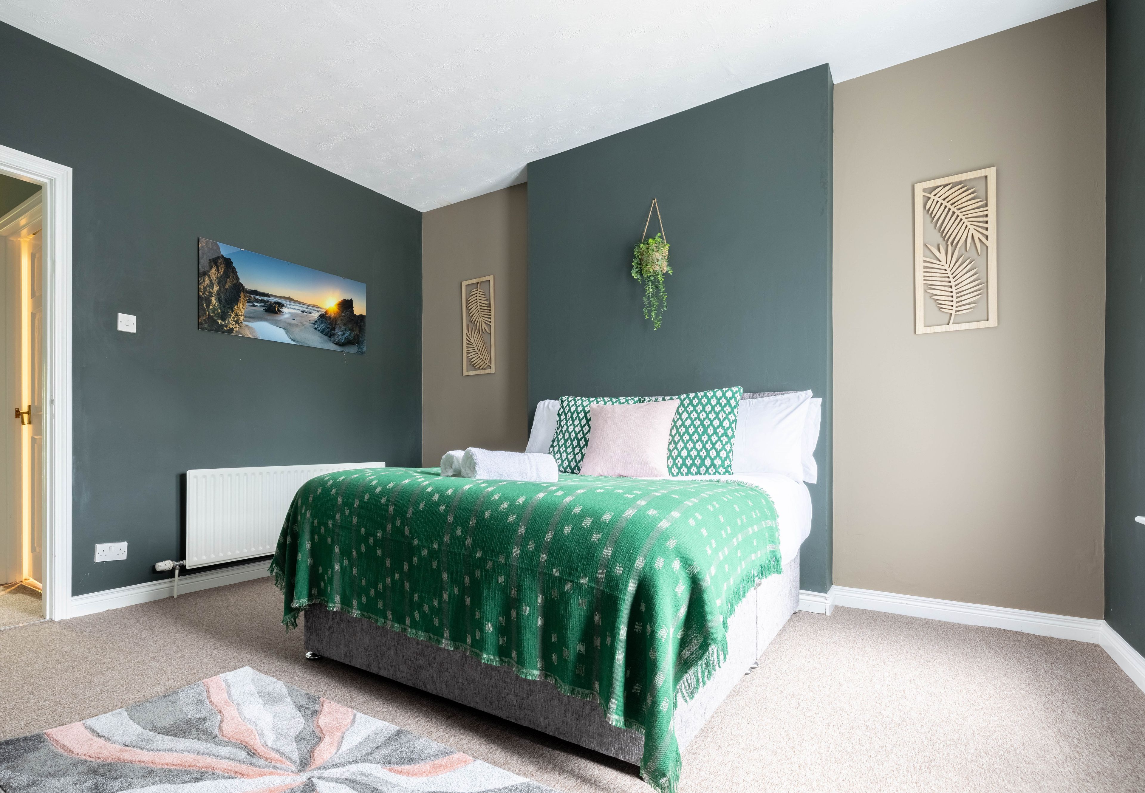 Quarto com cama, radiador, colcha estampada verde, pintura e planta pendente.