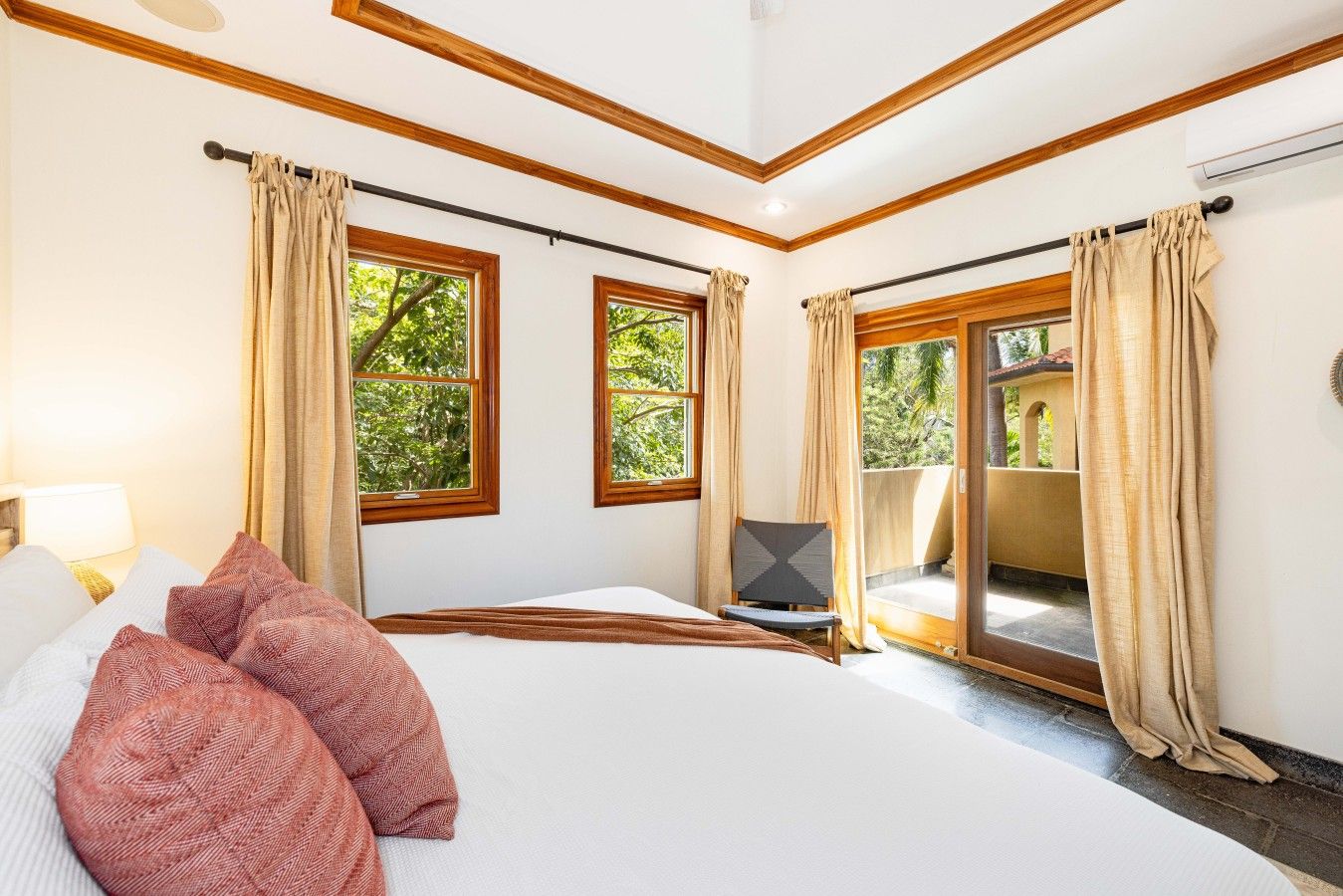 A hotel room with a large bed, two windows with curtains, a chair, and a view of greenery outside.