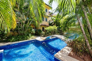 Une piscine à bord incurvé, entourée d’une végétation luxuriante et bordée d’un bâtiment à la façade jaune.