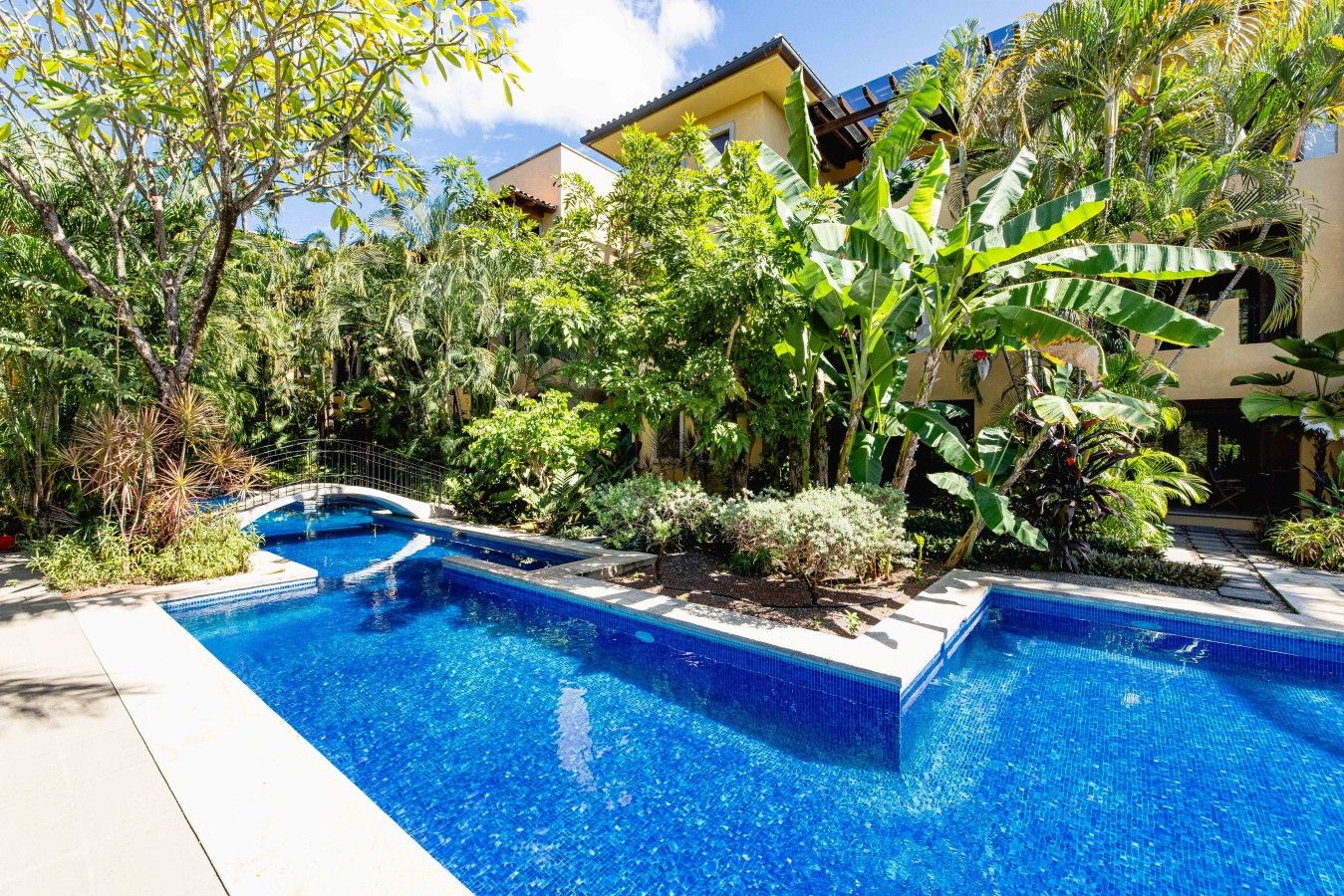 A swimming pool with a tiled edge, surrounded by lush tropical vegetation and a building with a tiled roof in the background.