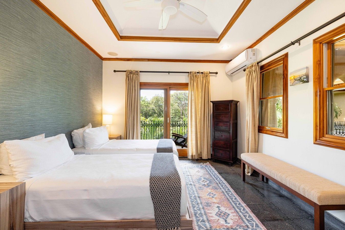 Une chambre d’hôtel avec deux lits, un banc, une grande fenêtre avec des rideaux, un ventilateur de plafond et une vue sur la verdure.