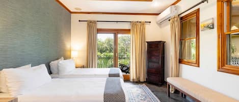 Een hotelkamer met twee bedden, een bankje, een groot raam met gordijnen, een plafondventilator en uitzicht op groen.