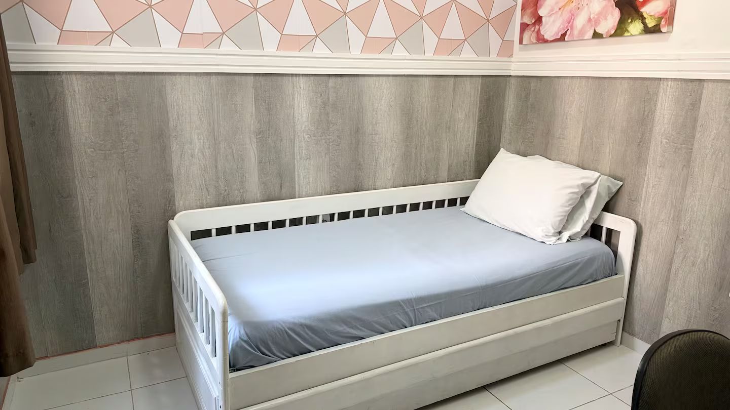 Ein Einzelbett mit weißem Gestell und grauem Lattenrost in einem Zimmer mit einer Holzwand und einer deckenhohen geometrischen Musterung.
