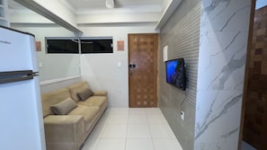 Un salon moderne avec un canapé beige, un téléviseur à écran plat fixé au mur et un réfrigérateur placé dans un coin.