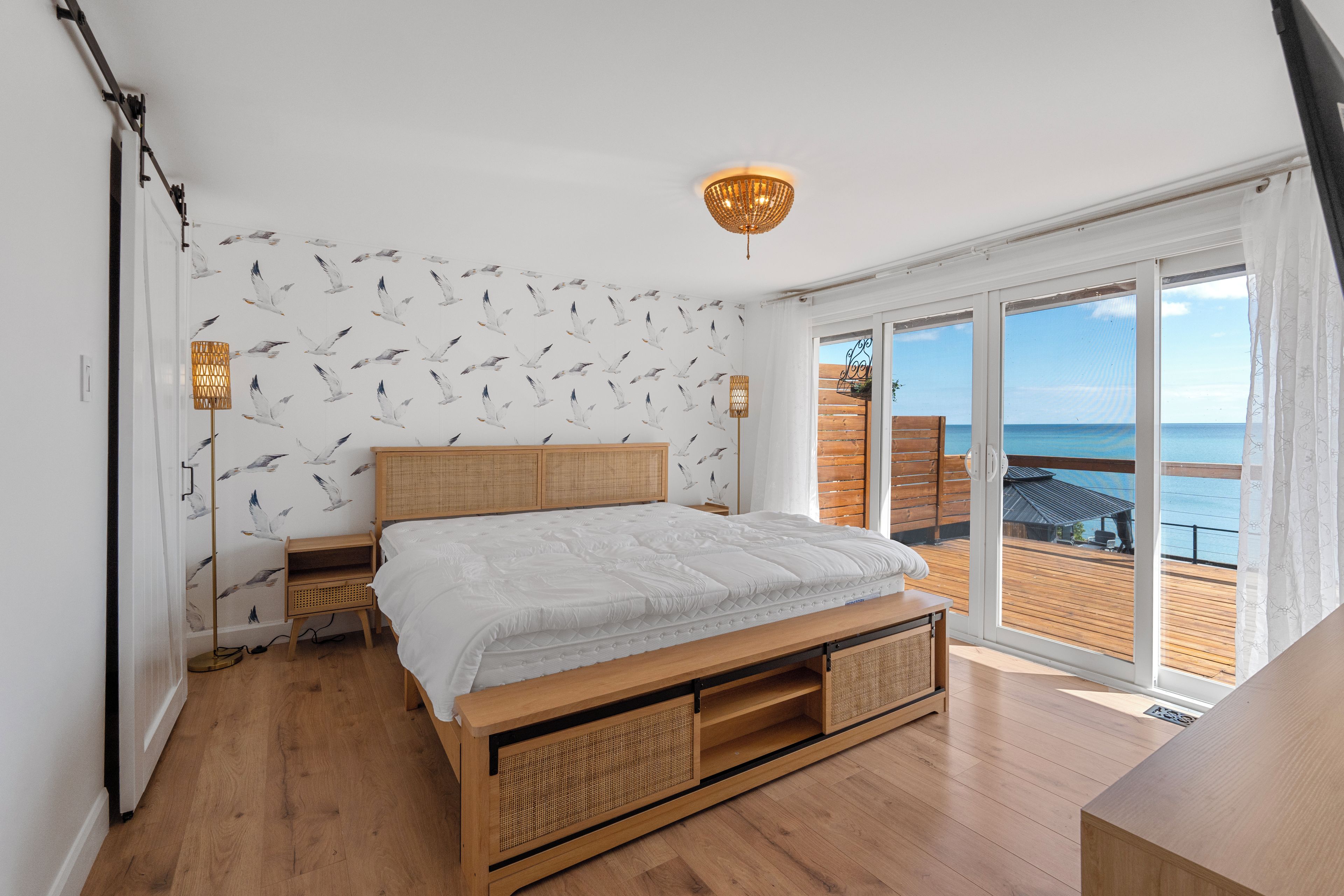 A bedroom with a large bed, wooden headboard, and a view of the sea through a sliding door.