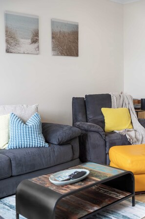A modern living room with a grey sofa, a yellow ottoman, and a coffee table with a magazine. There are two wall-mounted paintings above the sofa. - The Onka Contemporary Escape by the Coast (Port Noarlunga)