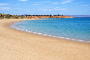A beach with a clear sky, sandy shore, and a distant coastline with buildings. - The Onka Contemporary Escape by the Coast (Port Noarlunga)
