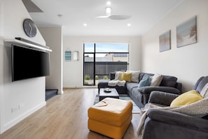 Ein modernes Wohnzimmer mit Flachbildfernseher, einem dunkelgrauen Sofa, einem gelben Hocker und einem großen Fenster mit Blick auf ein Gebäude mit Solaranlage.
