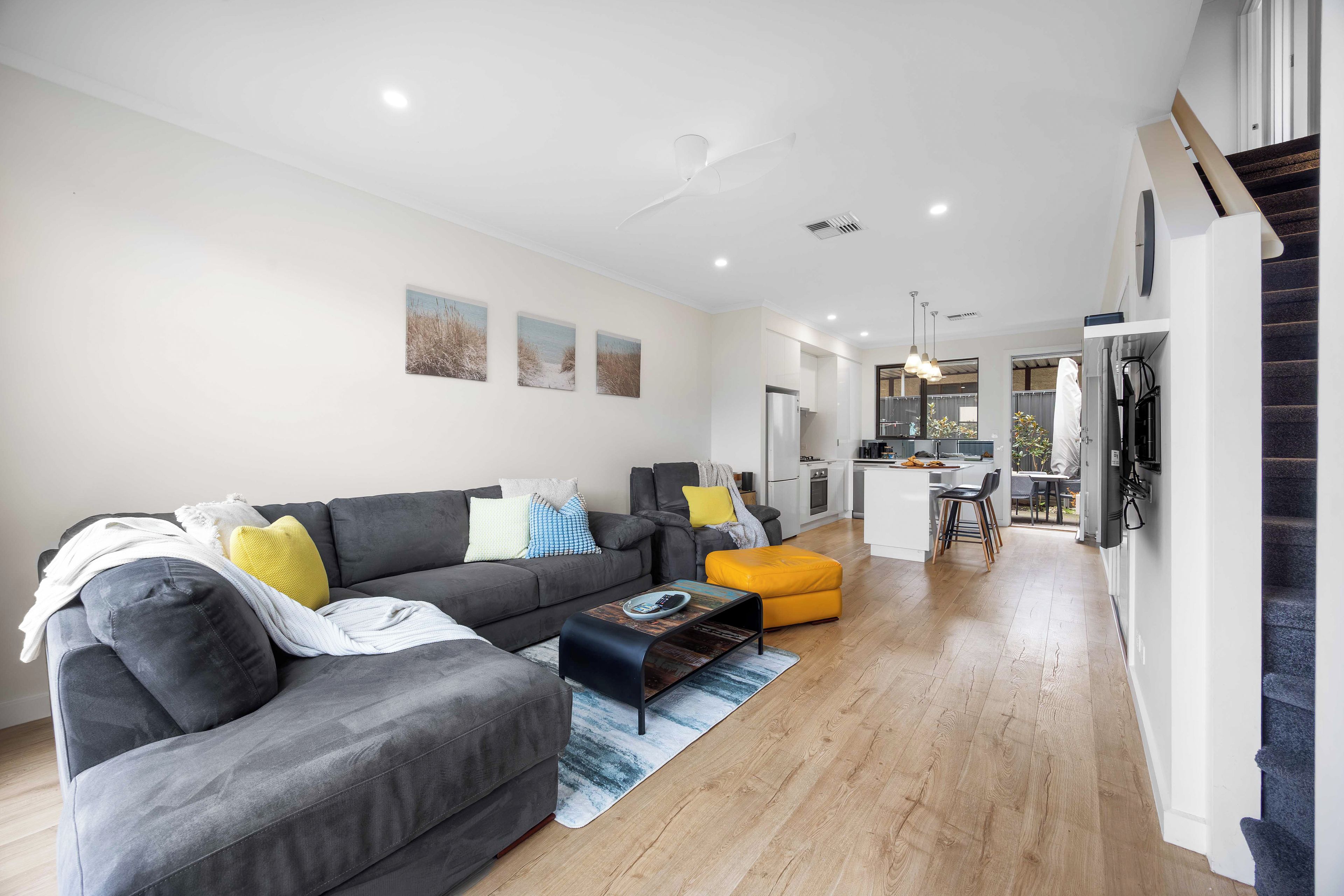 A modern living room with a grey sofa, a yellow ottoman, and a kitchen area in the background.