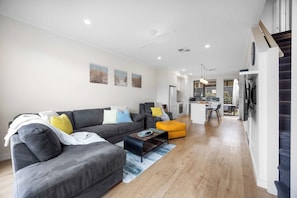 Ein modernes Wohnzimmer mit einem grauen Sofa, einem gelben Hocker und einer Küche im Hintergrund.