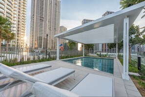 Ein moderner Poolbereich im Freien mit Liegestühlen, einer überdachten Lounge und Hochhäusern im Hintergrund.