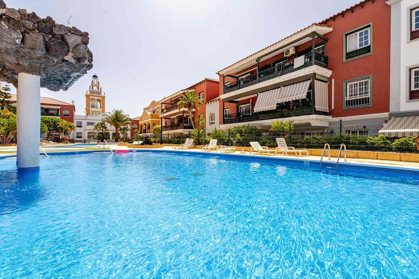 A swimming pool with lounge chairs, a stone structure, and buildings with balconies in the background.