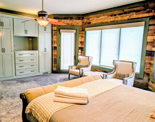 A bedroom with a large bed, two chairs, a desk with drawers, and a window with blinds.