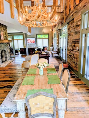 A spacious dining area with a long wooden table set for six, flanked by chairs with woven seat covers. A rustic chandelier hangs from the ceiling, and the room features exposed wooden beams and a stone fireplace.