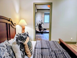 A bedroom with a bed, pillows, a lamp, and a view of a gym area.