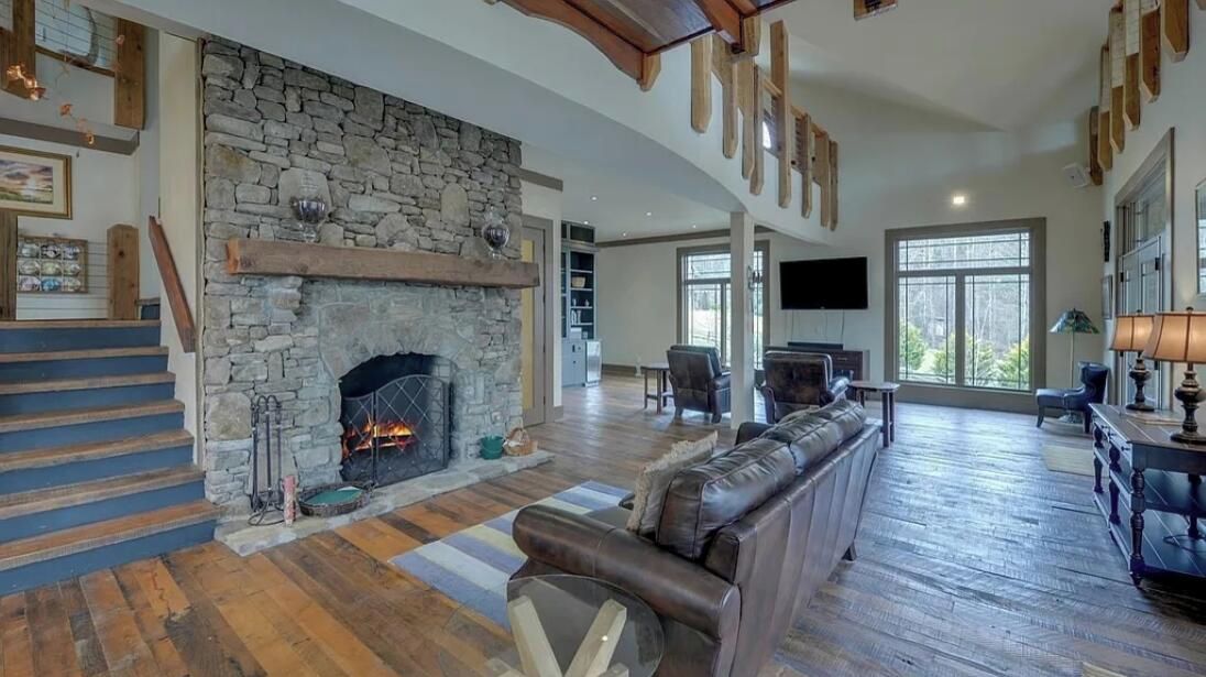 A spacious living room with a stone fireplace, a leather sofa, and a wooden staircase.