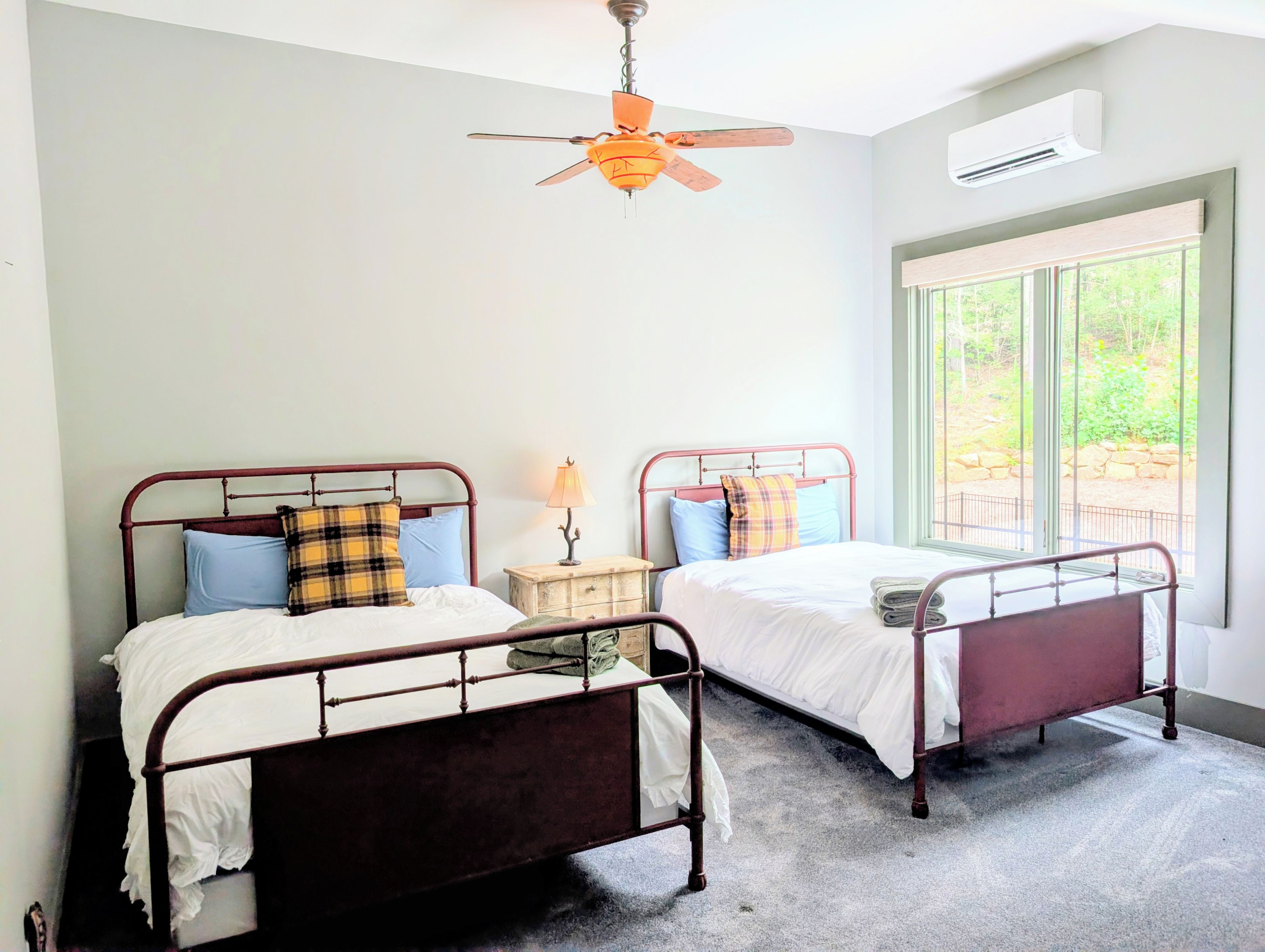 A bedroom with two metal-framed beds, a ceiling fan, an air conditioning unit, and a window with a view of greenery.