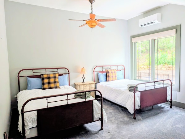A bedroom with two metal-framed beds, a ceiling fan, an air conditioning unit, and a window with a view of greenery.