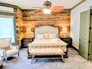 A bedroom with a tufted headboard, a bench at the foot of the bed, a chair, two bedside tables with lamps, and a wooden wall paneling.