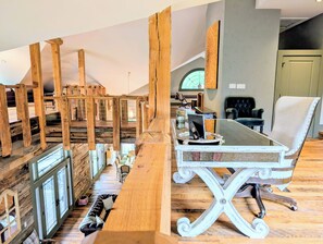 A room with a wooden staircase, a glass table with a laptop, a cushioned chair, and a window with a view.