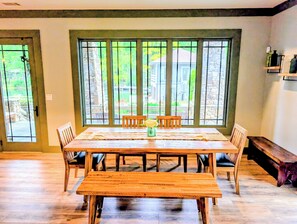 A dining area with a wooden table and chairs, large windows with a view of greenery, and a stone wall.