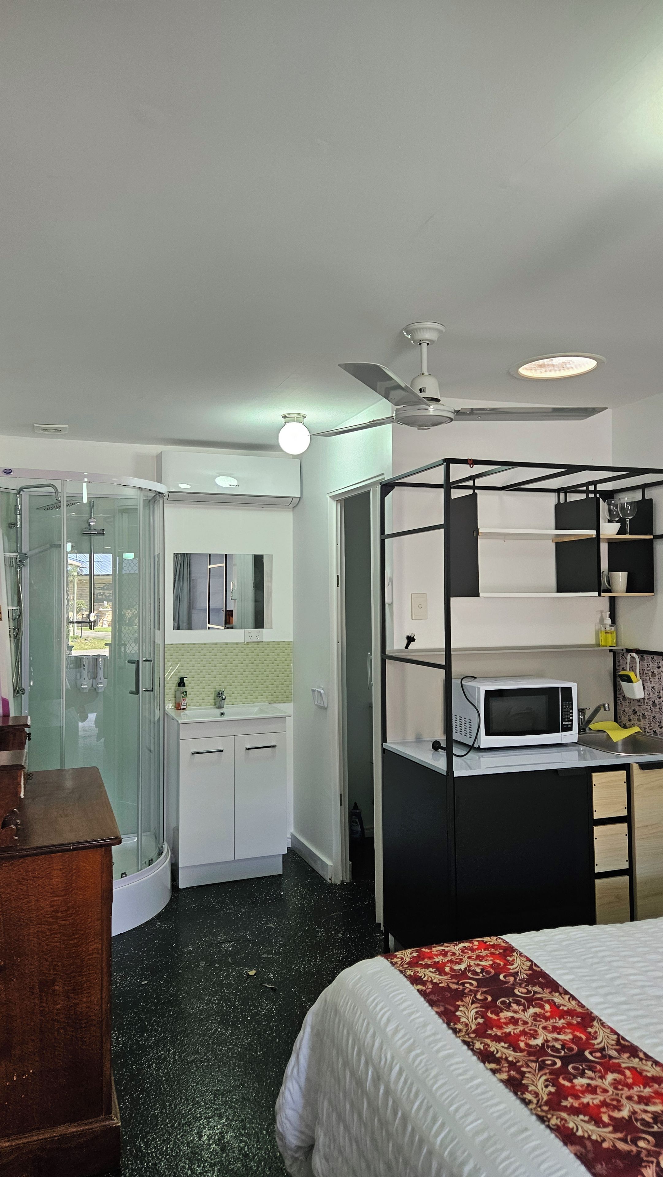 A hotel room with a bed, a kitchenette, a bathroom with a shower, and a ceiling fan.