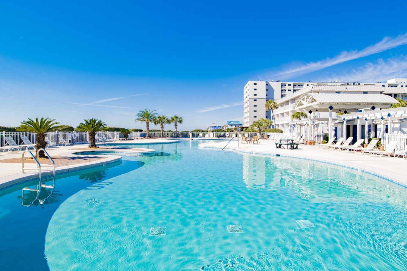 Una piscina azul cristalina con sillas de descanso y palmeras, rodeada por un edificio blanco con balcones.